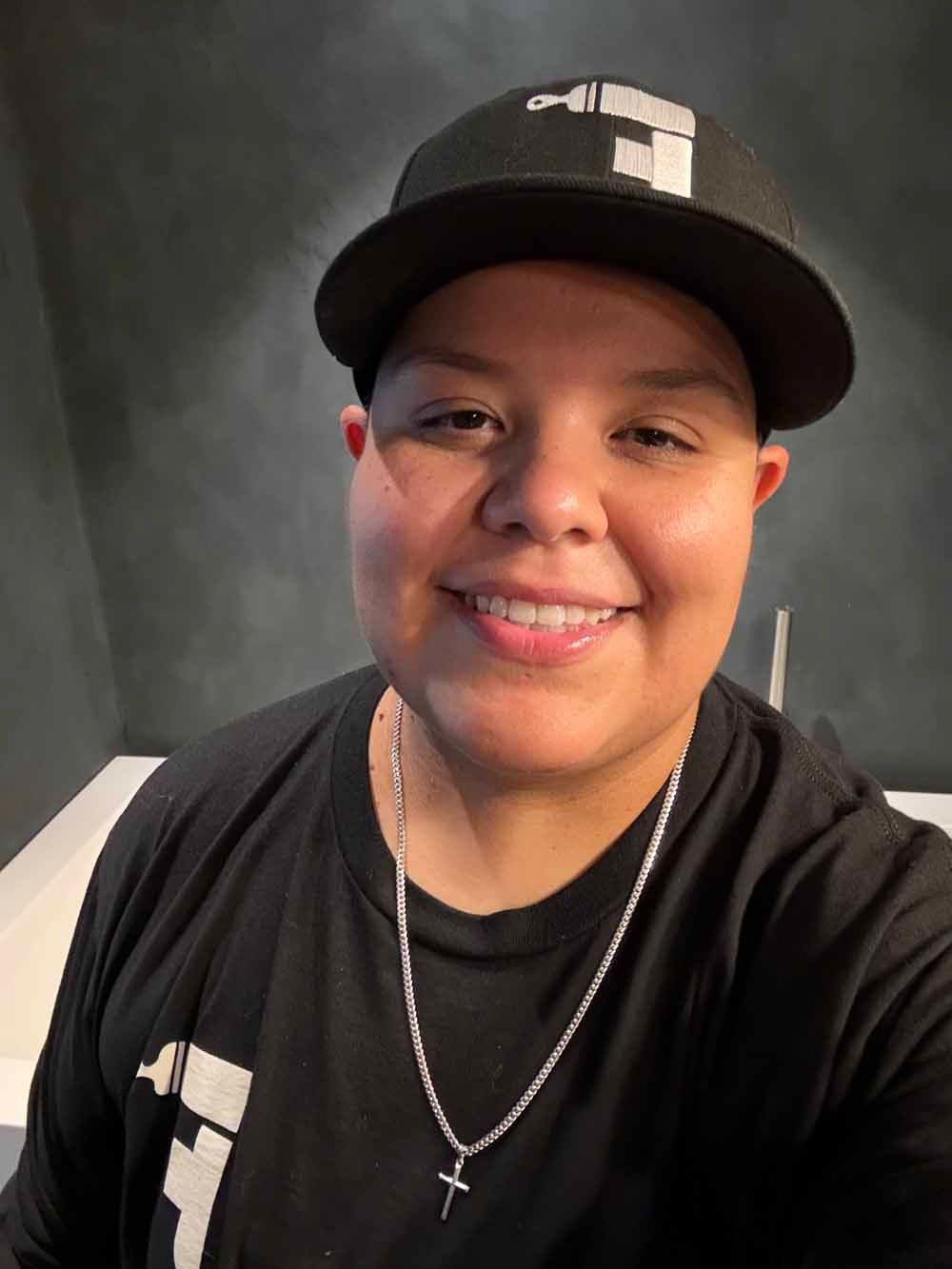 Person smiling in a black cap and shirt, wearing a silver cross necklace against a dark background.