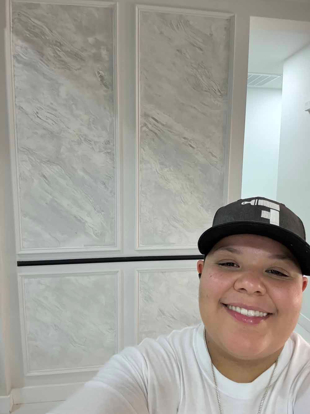 Smiling person in a black cap and white shirt taking a selfie indoors, with light marble-patterned walls behind.