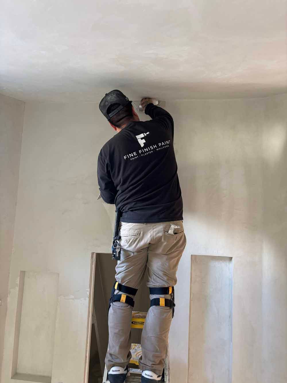 Worker plastering a ceiling indoors, wearing a black shirt and knee pads, on a step stool.