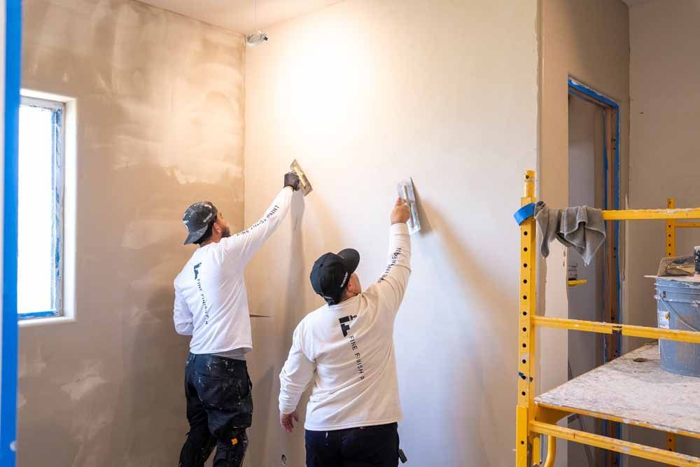 Two people painting a room’s white wall, with scaffolding and a window on the left.
