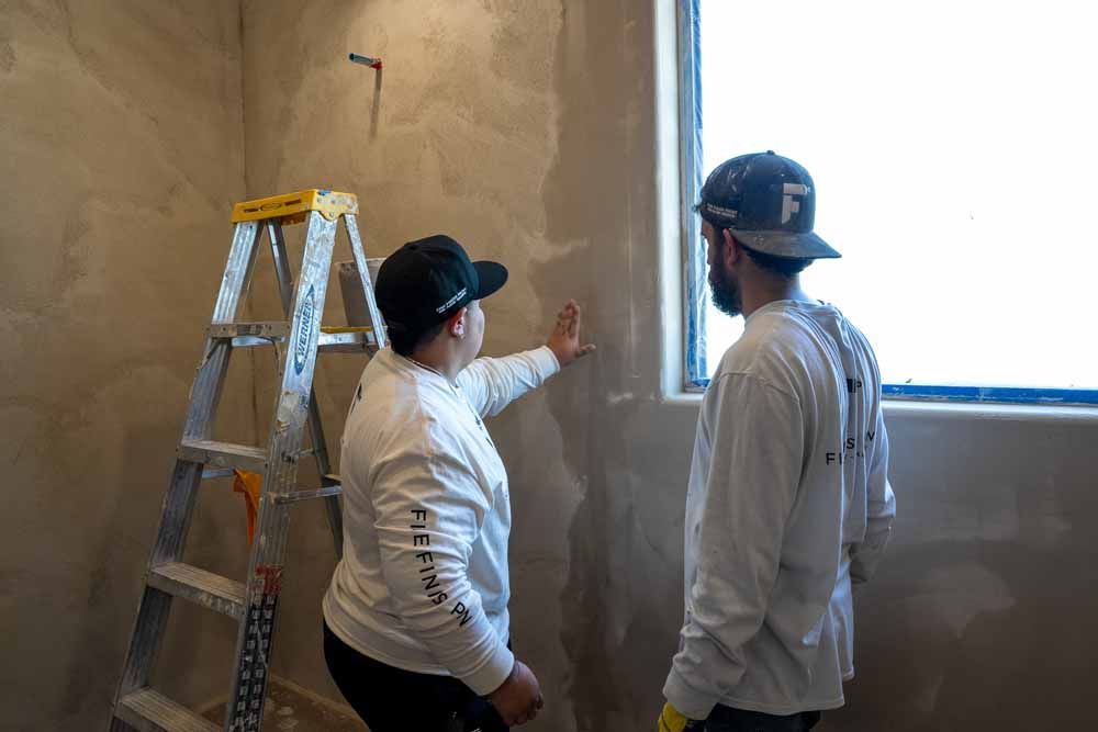 Two workers inspect a wall beside a ladder and window opening in an unfinished room.