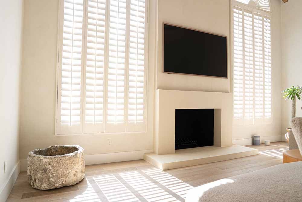 Bright living room with white shutters, a black wall-mounted TV above a fireplace, and sunlight on the floor