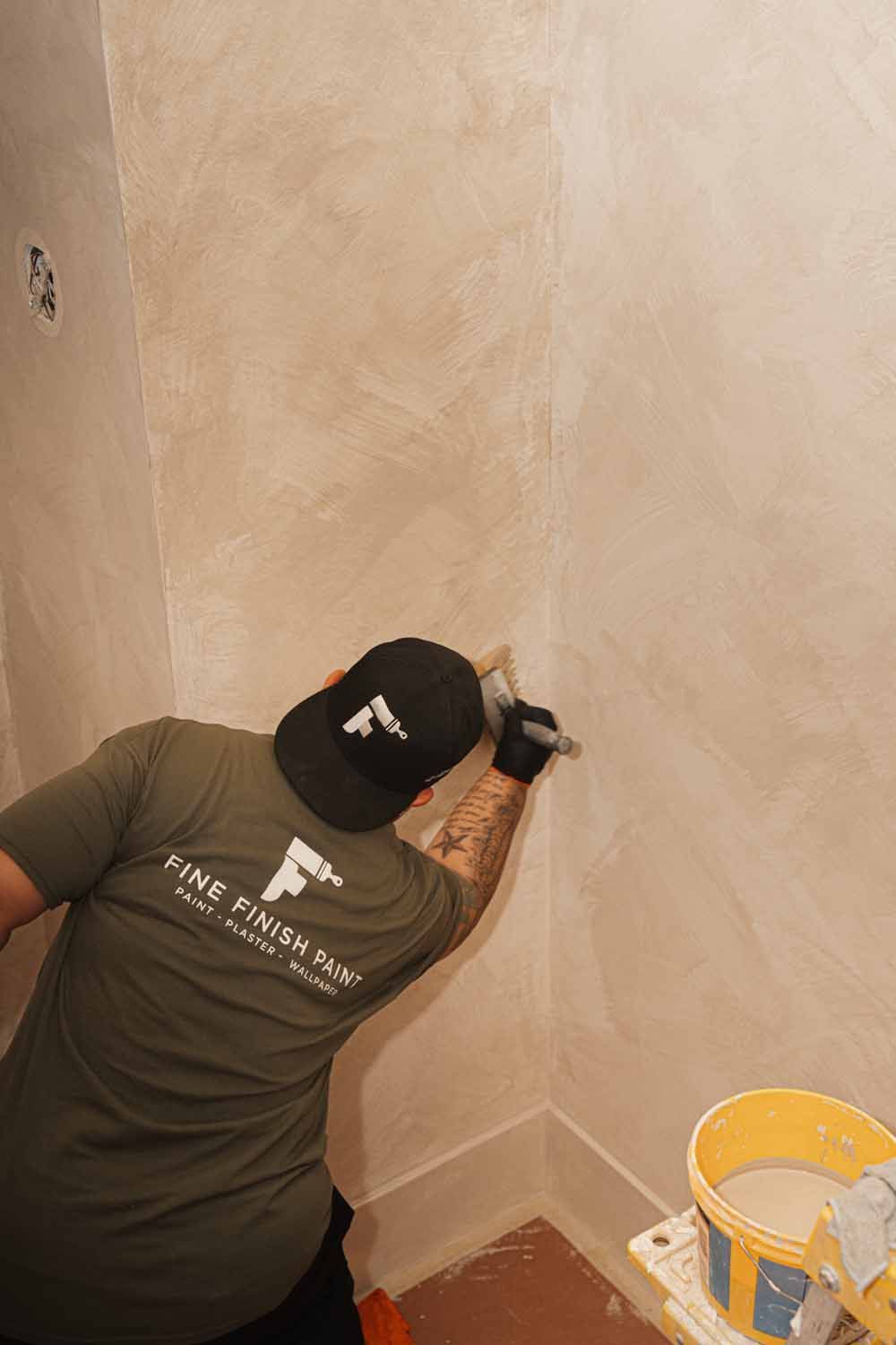Person painting a beige wall in a corner, with a yellow paint bucket on the floor.