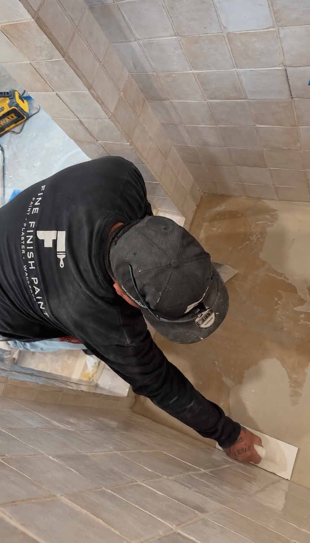 Person in black hoodie cleaning beige tiled wall with a cloth in a bathroom or shower area.