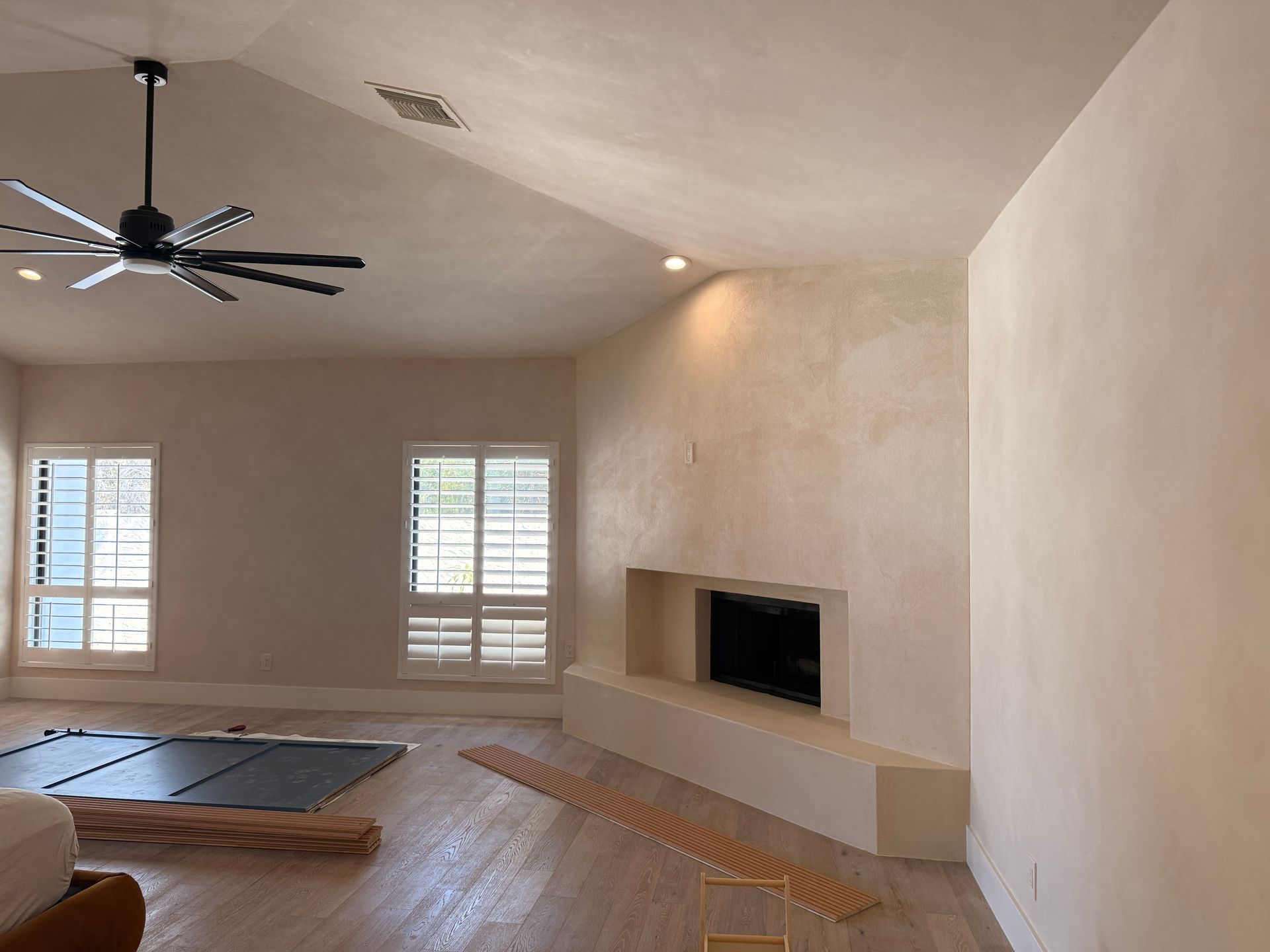 Spacious living room with beige walls, fireplace, wood floor, ceiling fan, and bright windows with shutters