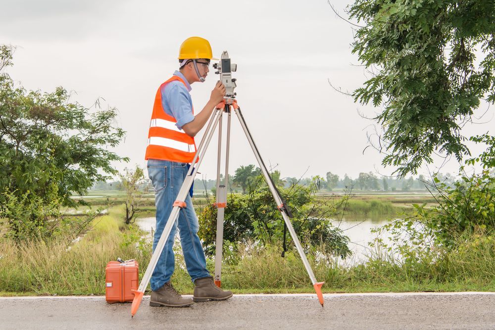 Surveyor in orange safety vest and yellow hard hat using a tripod instrument by a roadside