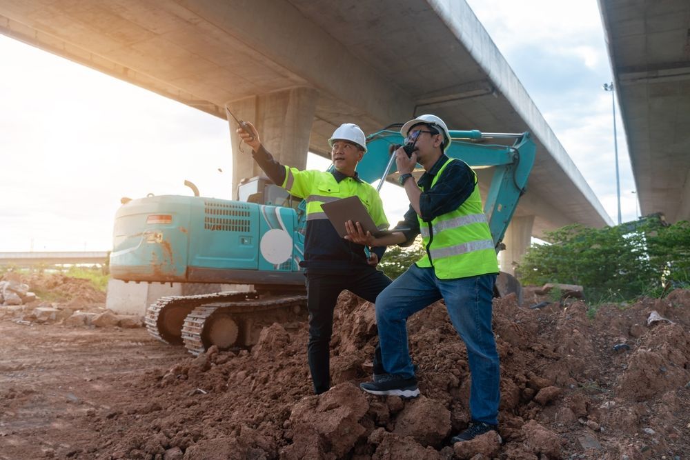 Two construction workers in safety vests and hard hats beside an excavator under an overpass.