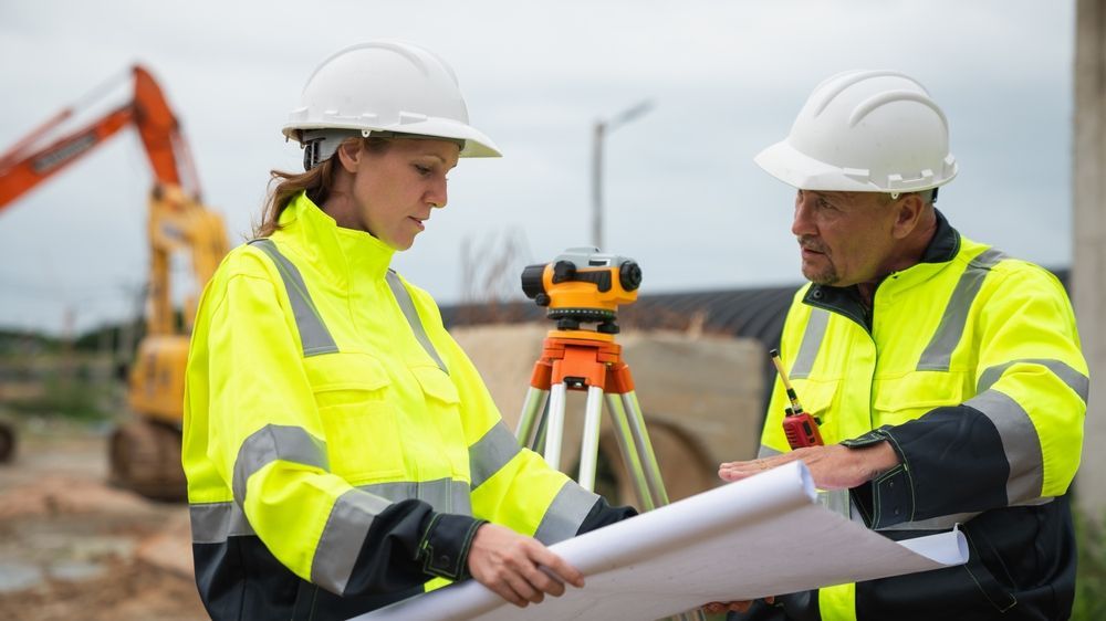 Two construction workers in hard hats and neon jackets review plans beside survey equipment at a worksite