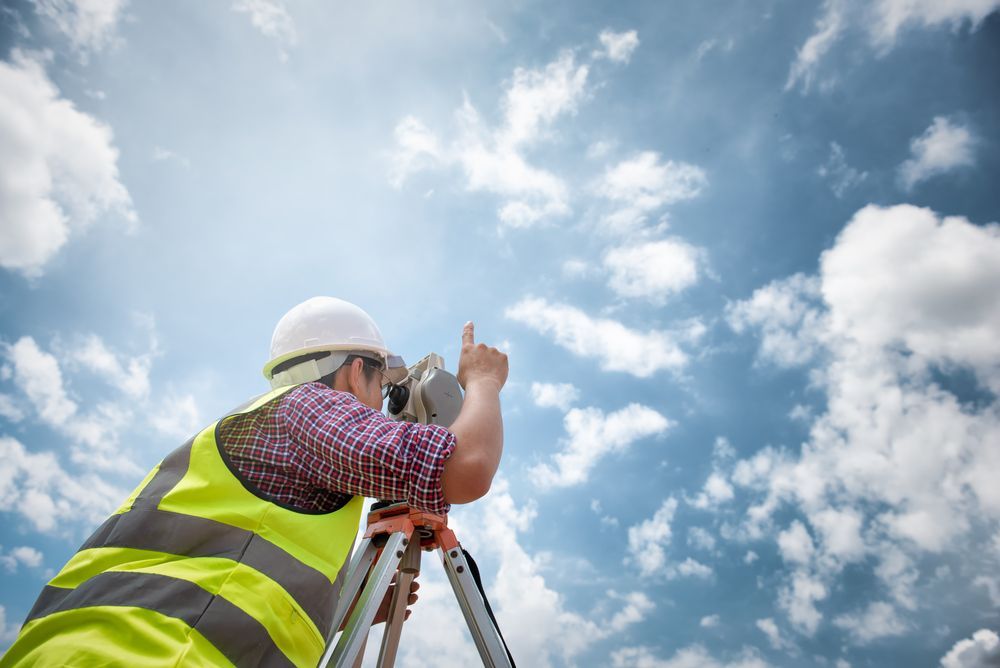 Surveyor in hard hat and safety vest using a tripod against a cloudy blue sky