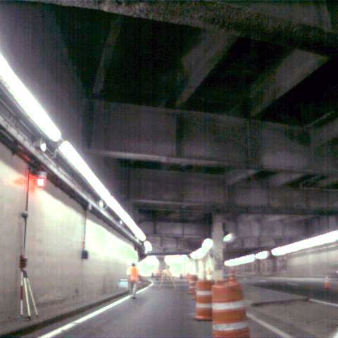 Inside a dim tunnel, a worker stands near orange traffic barrels under concrete beams.