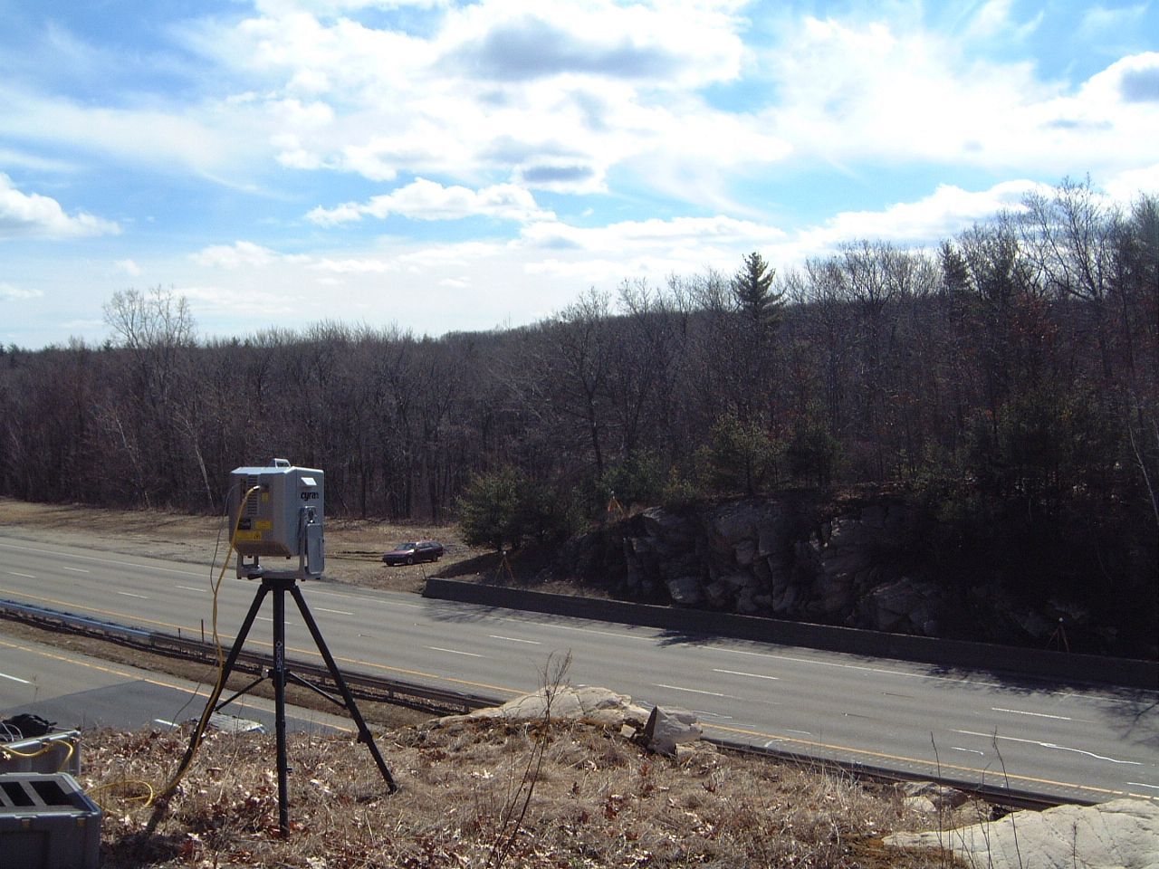 Surveying tripod with instrument beside a road under a partly cloudy sky, backed by leafless trees