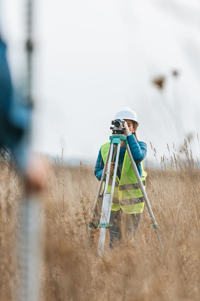 Surveyor in hard hat and safety vest using a tripod-mounted instrument in a grassy field