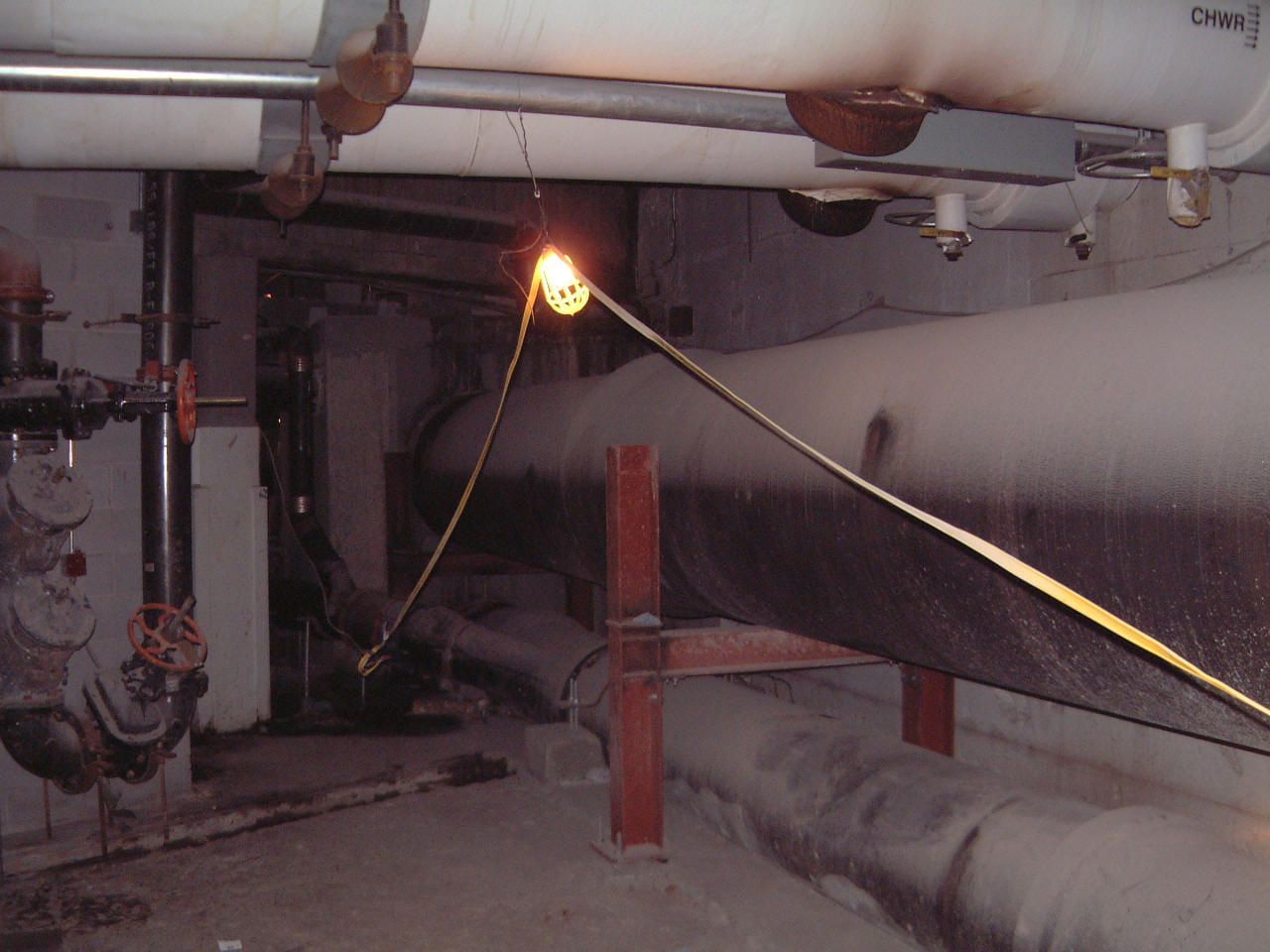 Dim industrial basement with pipes, a small flame, and a person using a long pole.