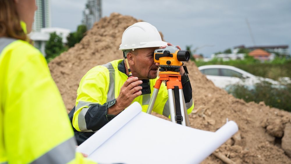 Surveyor in a white hard hat using a tripod instrument at a construction site with dirt piles and blueprints.