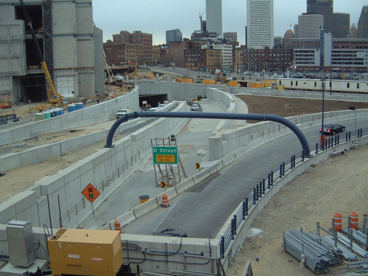Road construction site with concrete barriers, a curved ramp, and city buildings in the background.