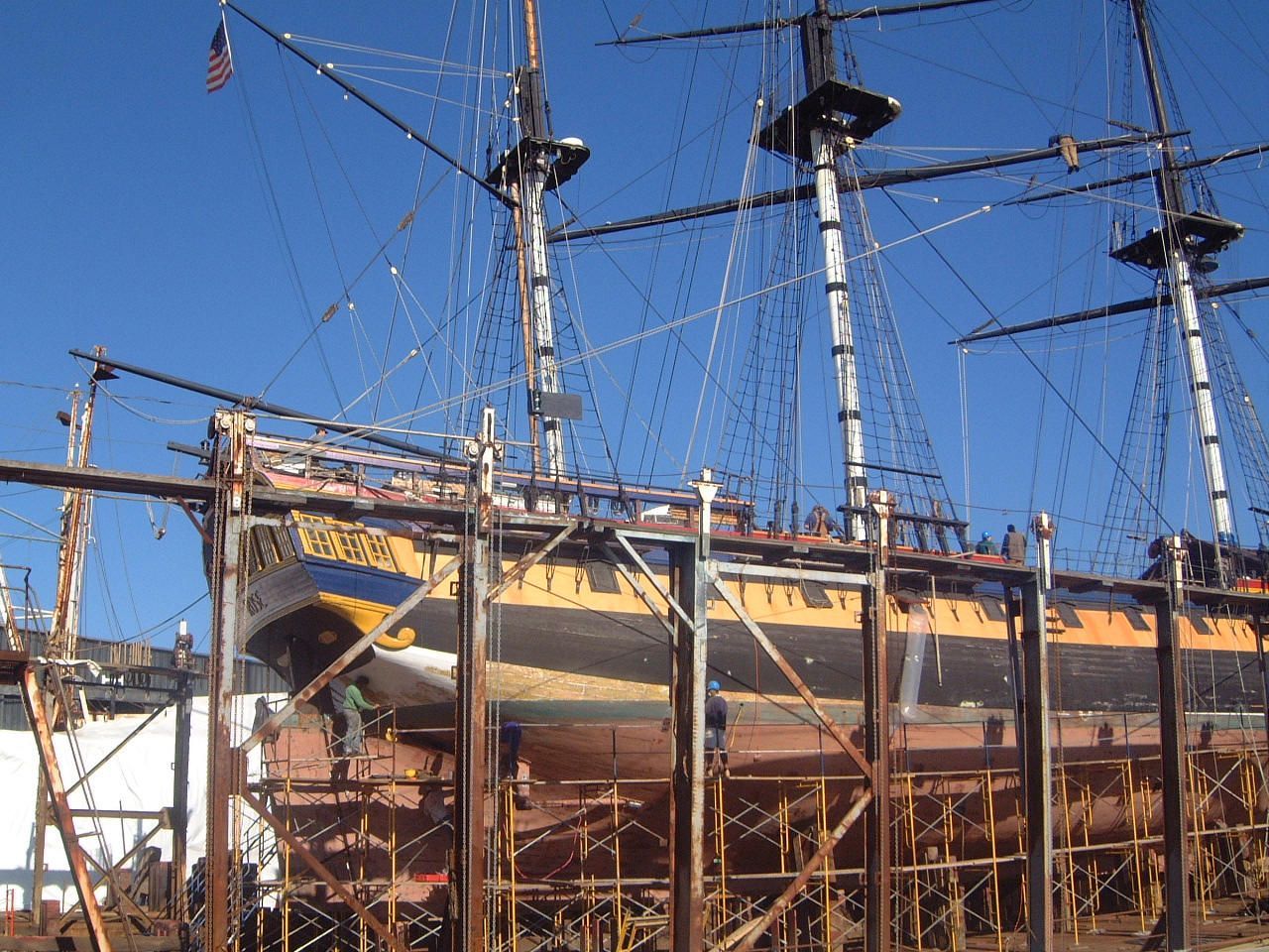 Historic wooden ship under restoration, with tall masts, rigging, and scaffolding against a blue sky
