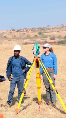Two surveyors in hard hats standing by a theodolite on a tripod in a dry field