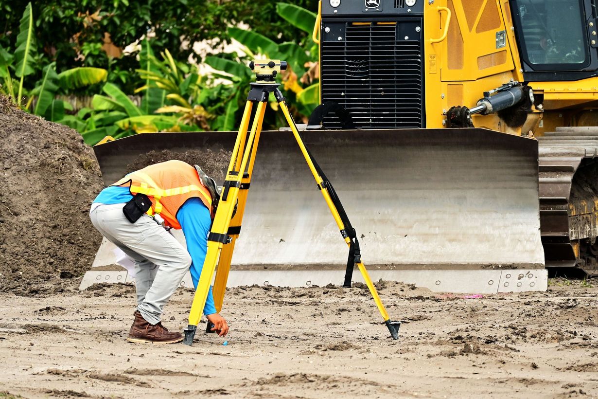 Surveyor setting up tripod equipment on muddy construction site beside a yellow bulldozer