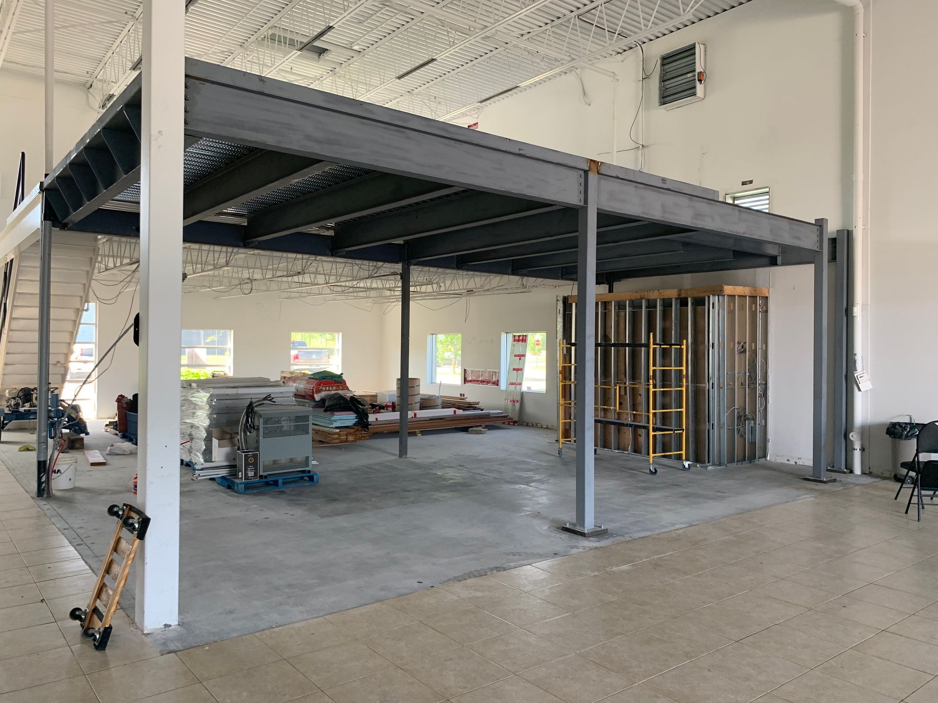 Vue intérieure d'un bâtiment avec une mezzanine métallique en construction. On aperçoit le sol en béton, les murs blancs et la charpente.