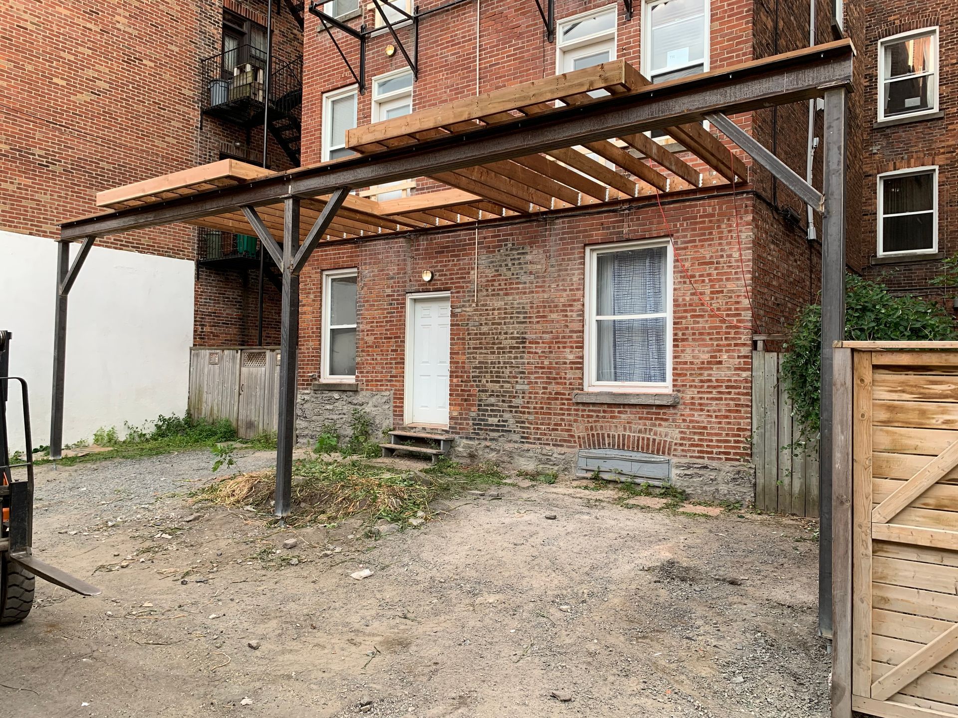 Un bâtiment en briques avec une pergola métallique. Une cour gravillonnée devant.