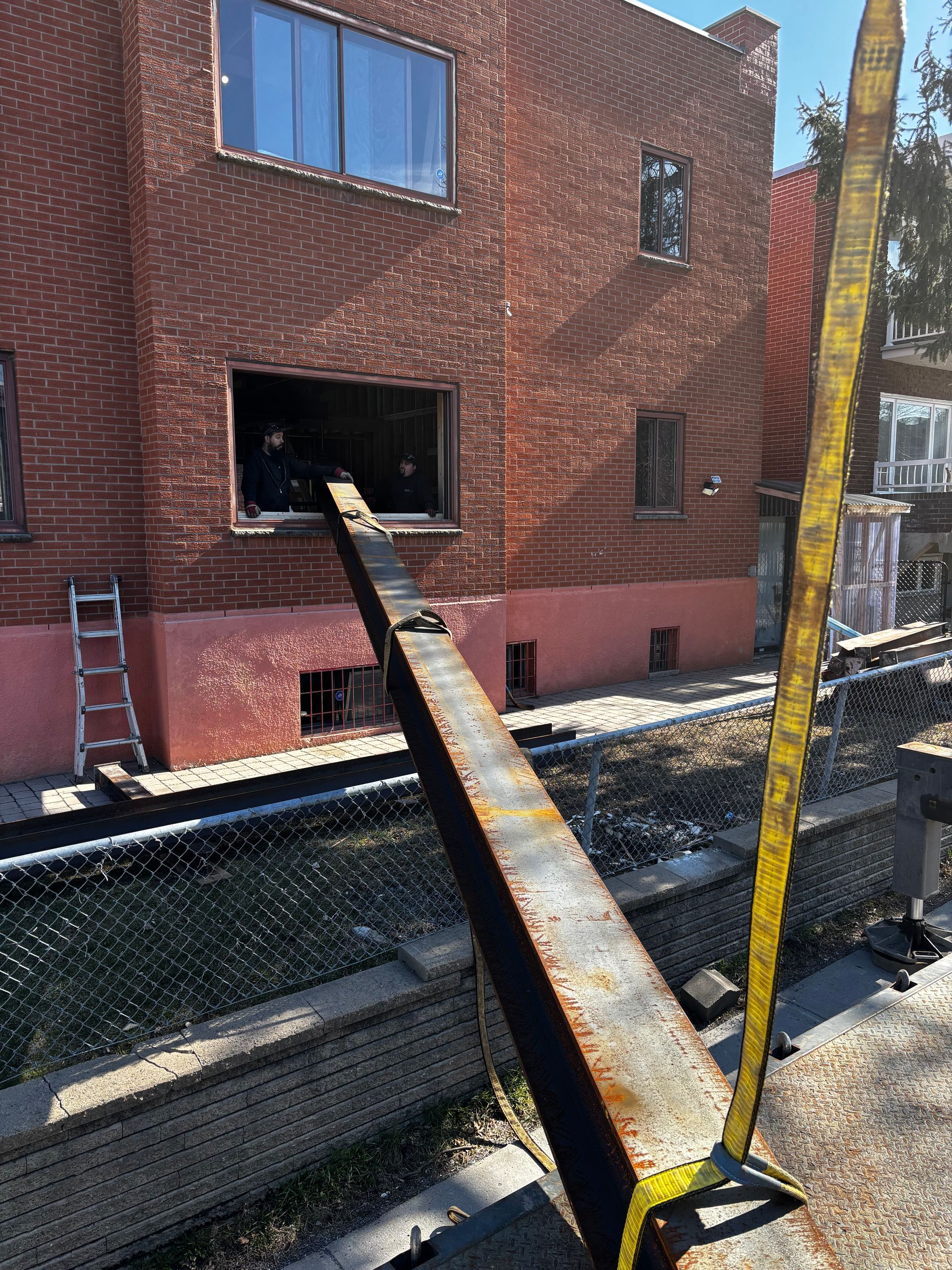 Une poutre métallique prolonge la façade d'un bâtiment. Une sangle jaune soutient la poutre ; une échelle se trouve à côté du bâtiment et une clôture est visible au premier plan.