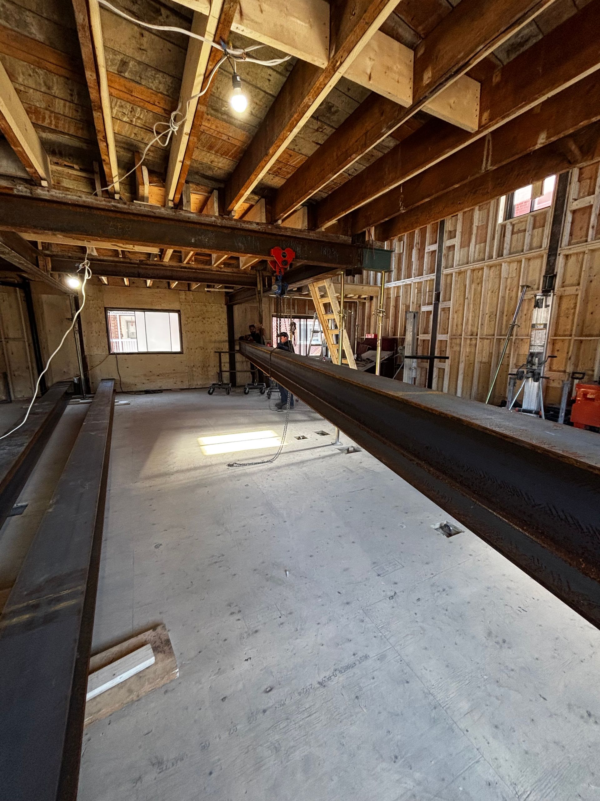 Intérieur d'un chantier de construction : sol en béton, poutres en bois apparentes, poutres en acier et ossature murale partielle.