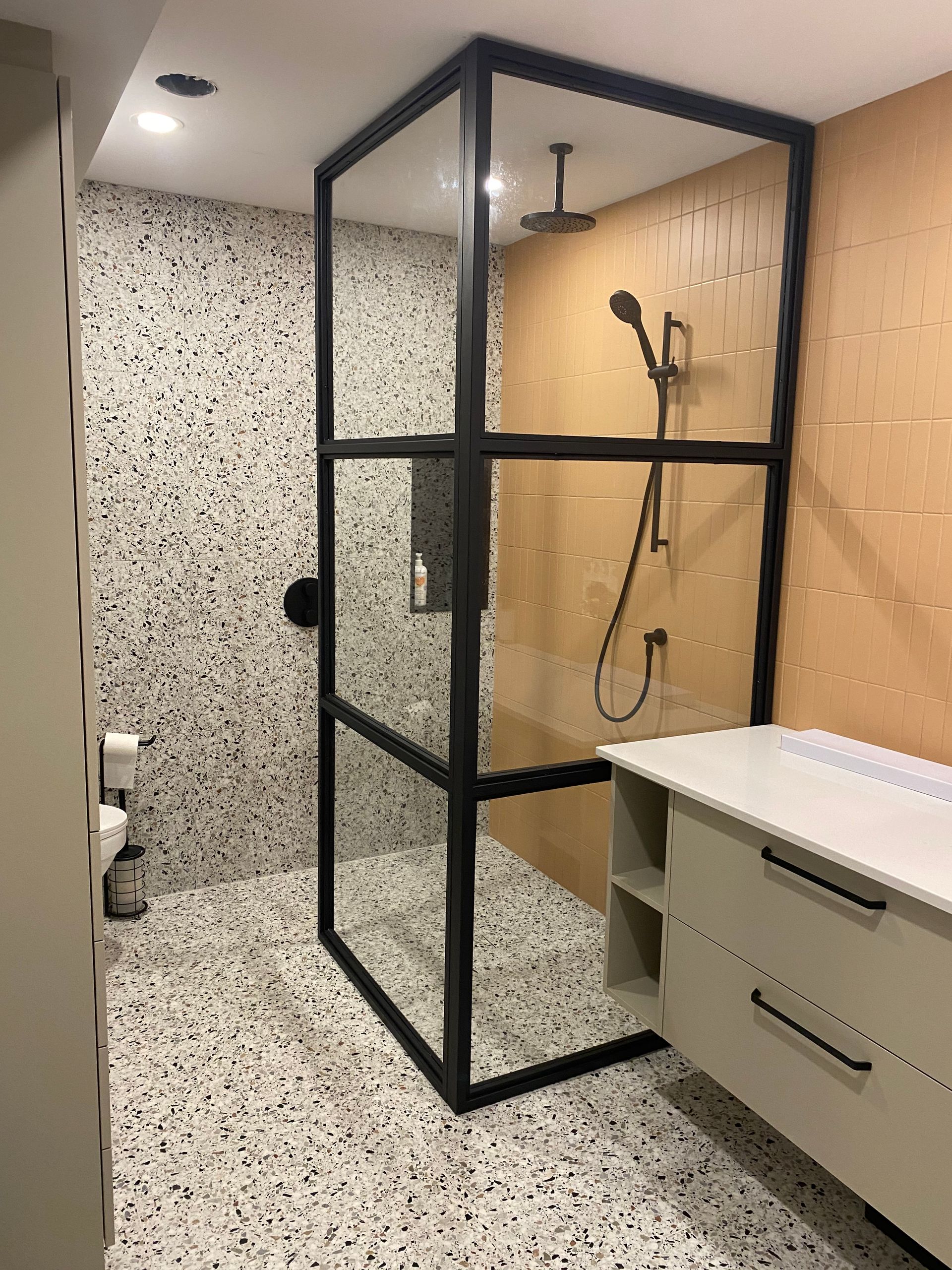 Salle de bains moderne avec douche à paroi de verre encadrée de noir, sol en carrelage terrazzo, murs beiges et meuble-lavabo de couleur claire.