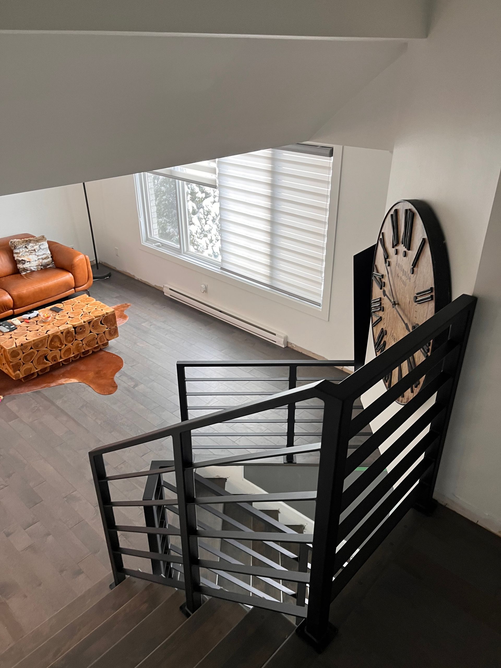 Vue depuis l'escalier : Intérieur moderne avec parquet, rampe noire, horloge surdimensionnée, canapé en cuir et stores.