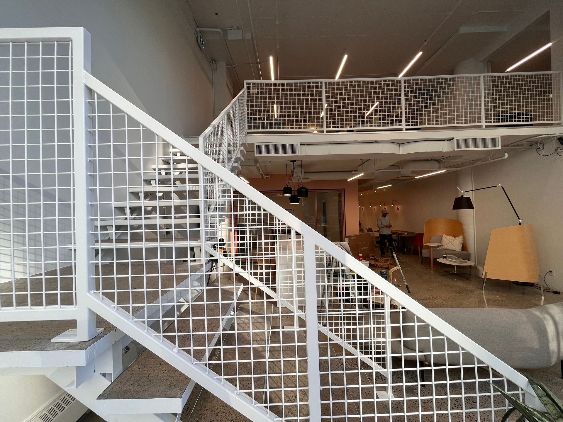Un escalier en métal blanc mène à une mezzanine. Vue intérieure : espace ouvert, mobilier moderne, une personne est visible.