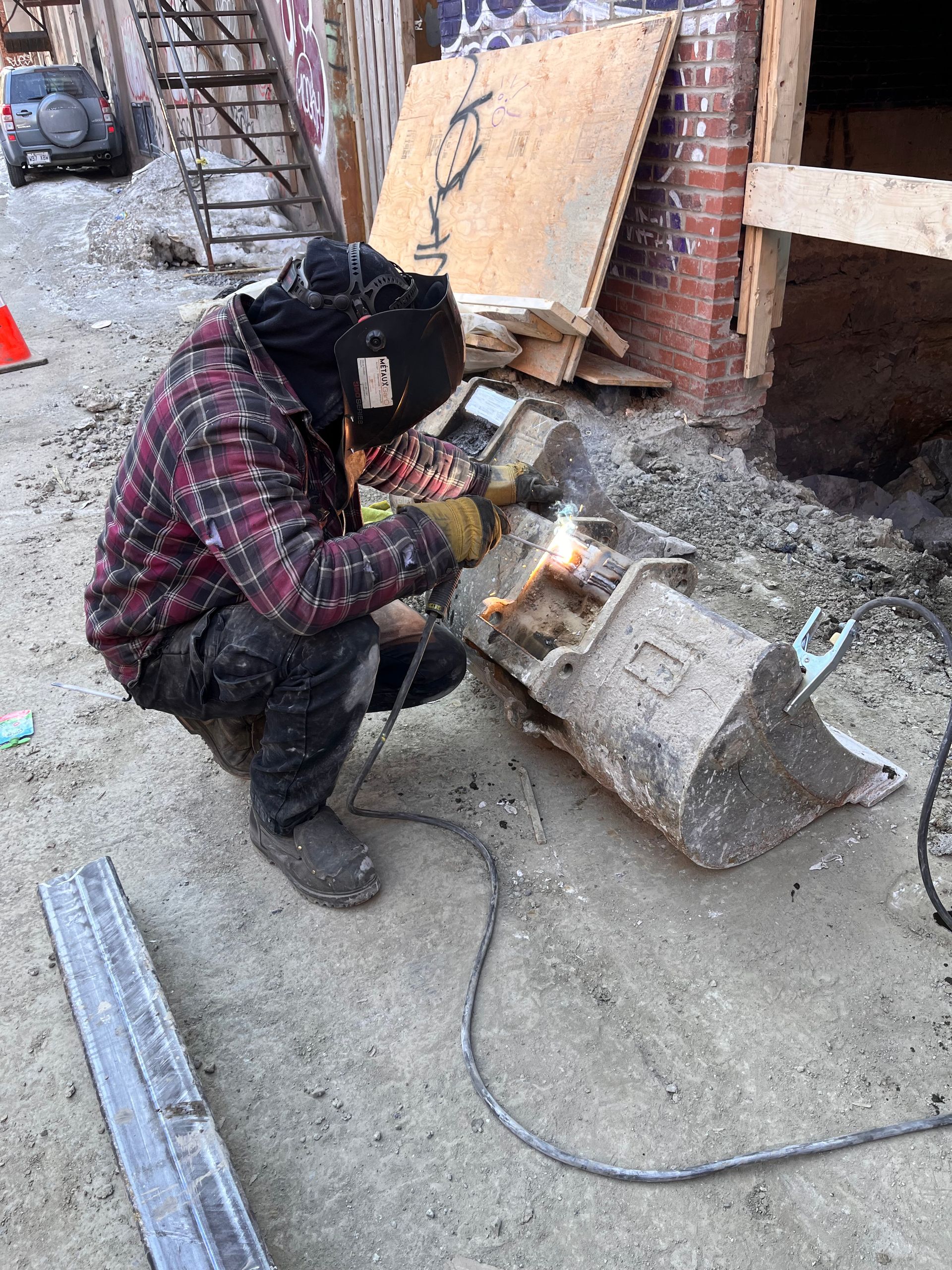 Une personne portant un équipement de soudage soude un seau métallique sur un chantier extérieur.