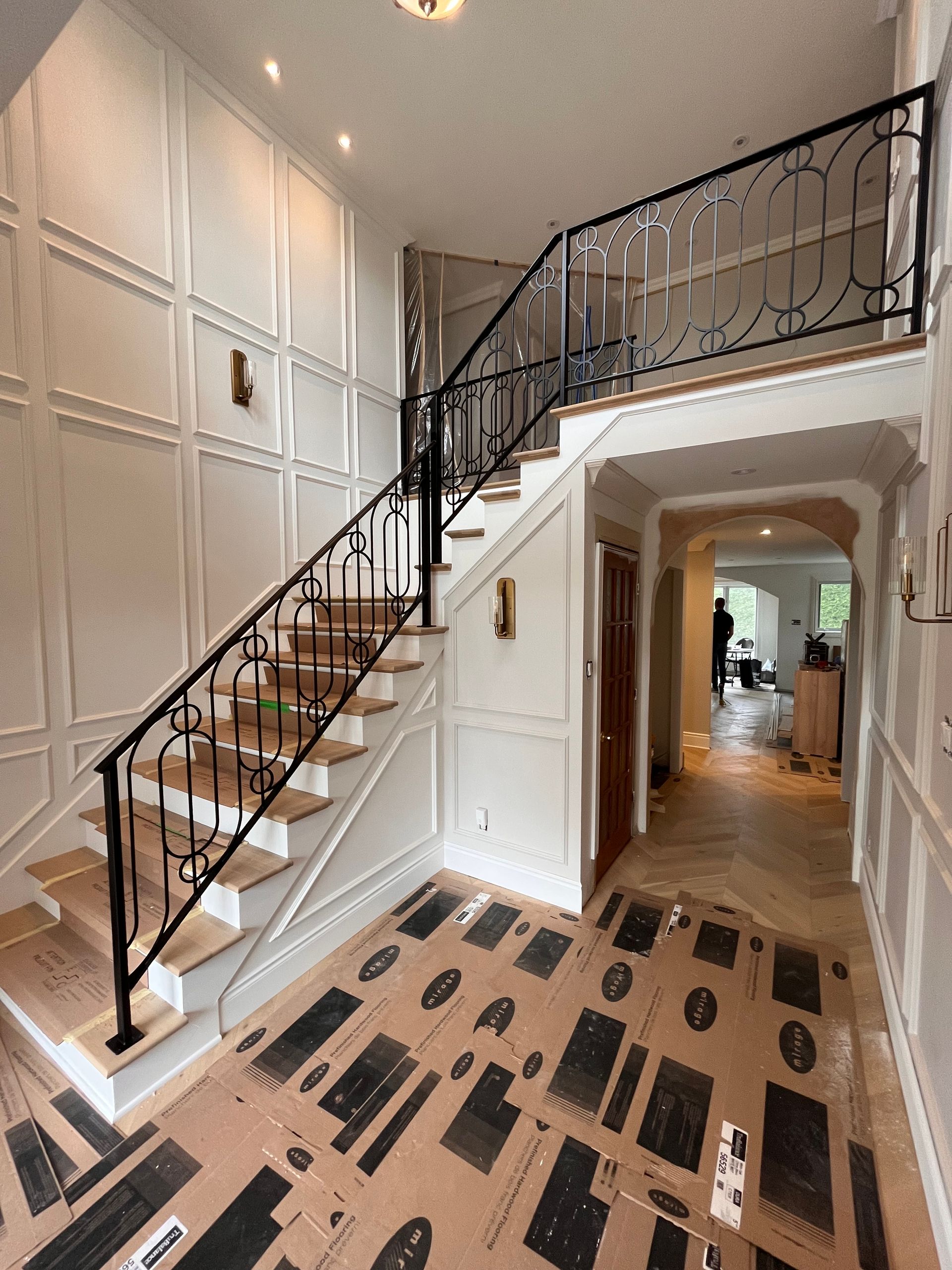 Escalier dans une maison avec rampe noire, lambris blancs et parquet recouvert de papier protecteur.