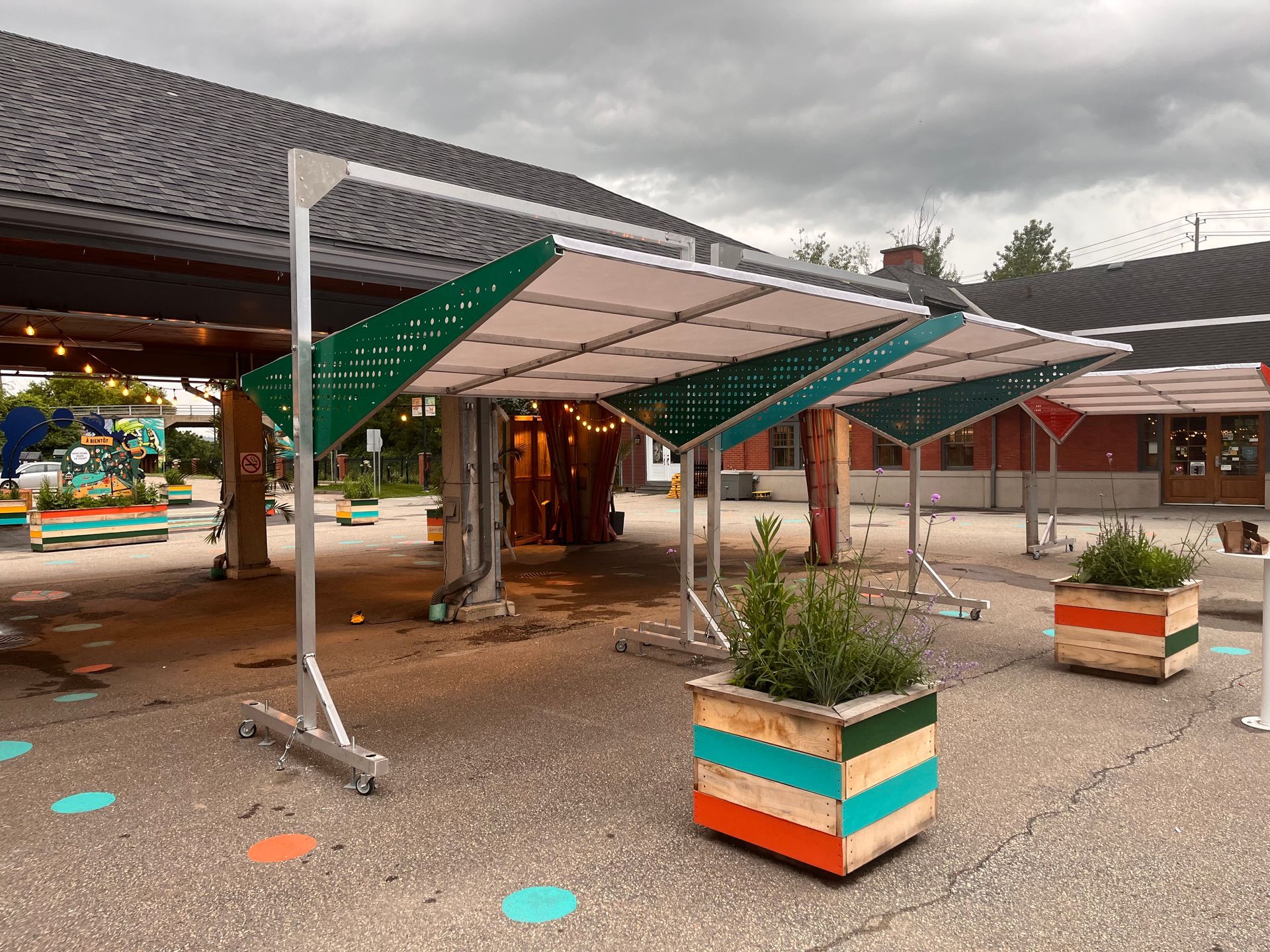 Une structure en forme de canopée verte et blanche, soutenue par des cadres métalliques, surplombe un patio agrémenté de jardinières colorées.