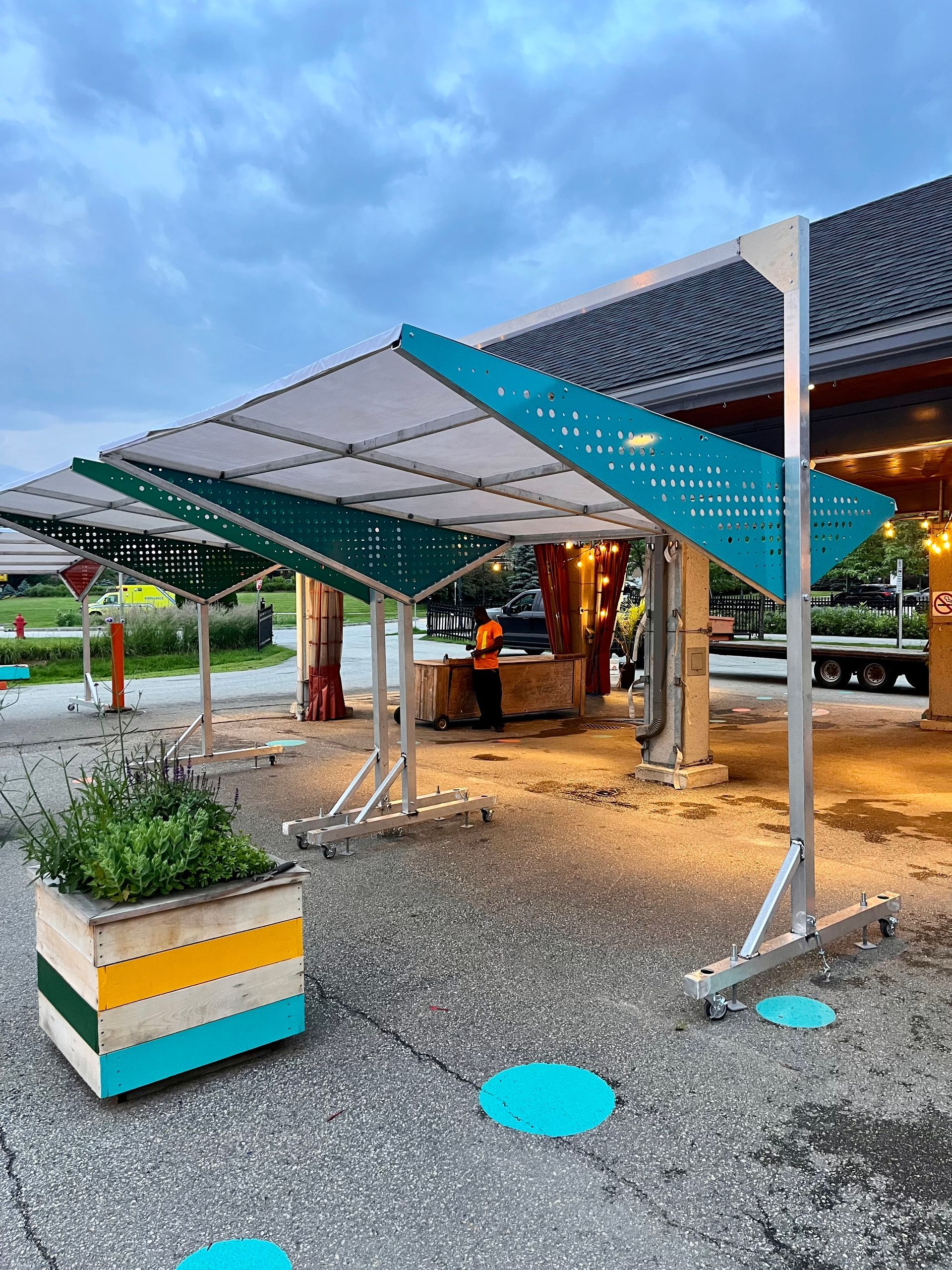 Espace extérieur aménagé avec une canopée turquoise et blanche, un sol en béton, des jardinières et des repères au sol bleus.