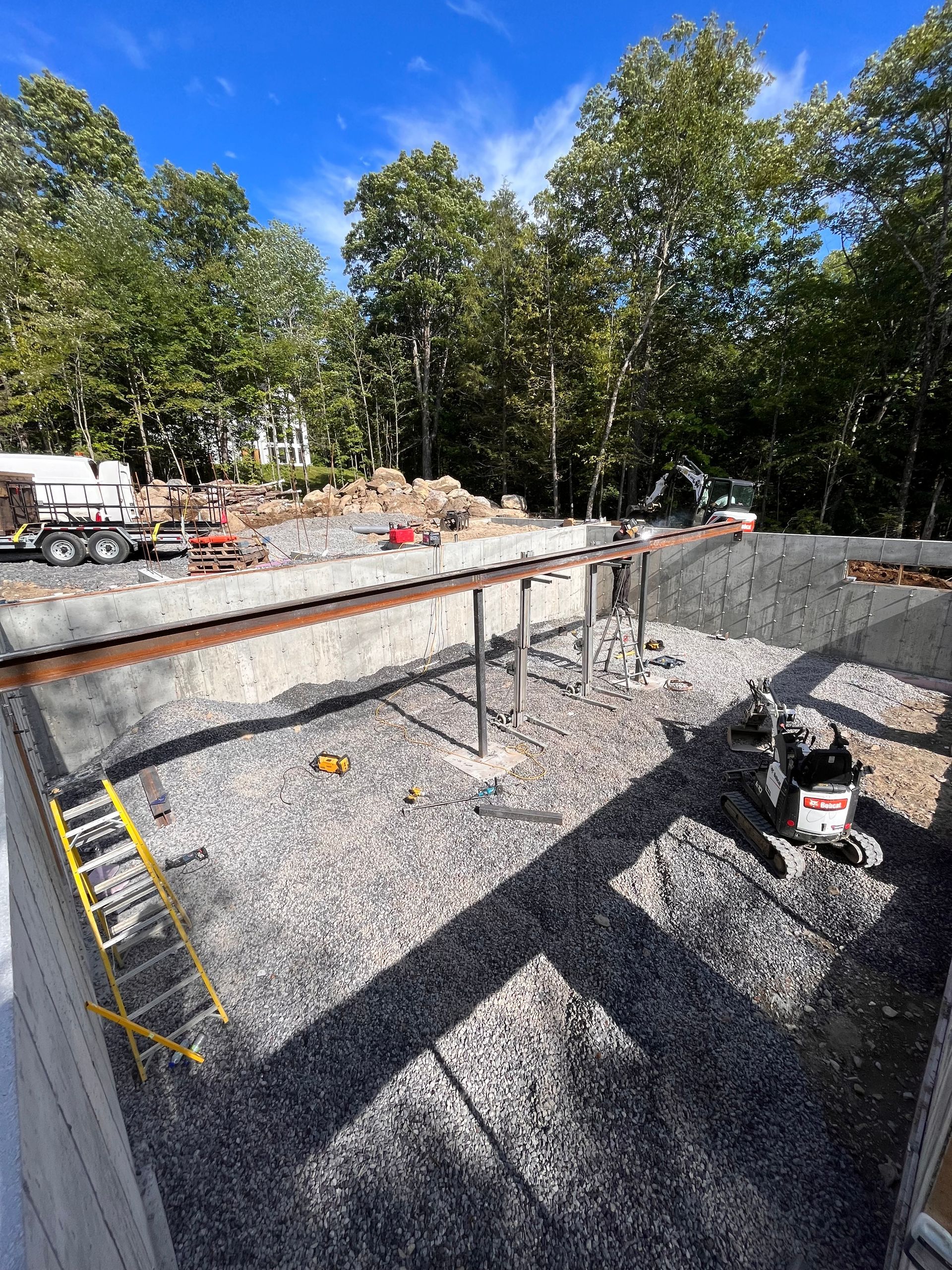 Chantier de construction avec gravier, poutres, échelle et matériel ; arbres en arrière-plan.