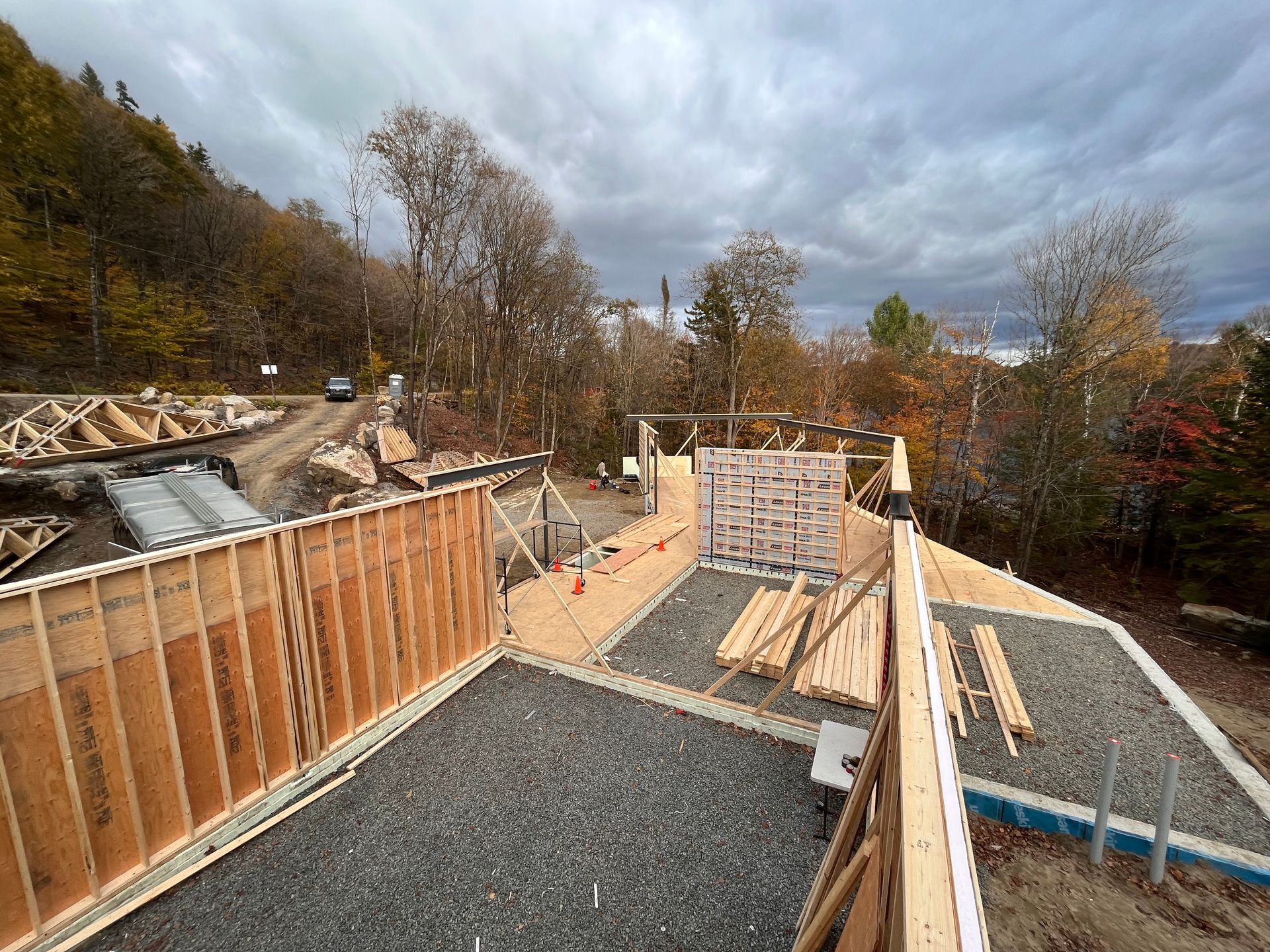 Chantier de construction avec charpente en bois et gravier, sous un ciel nuageux. Arbres et route en arrière-plan.