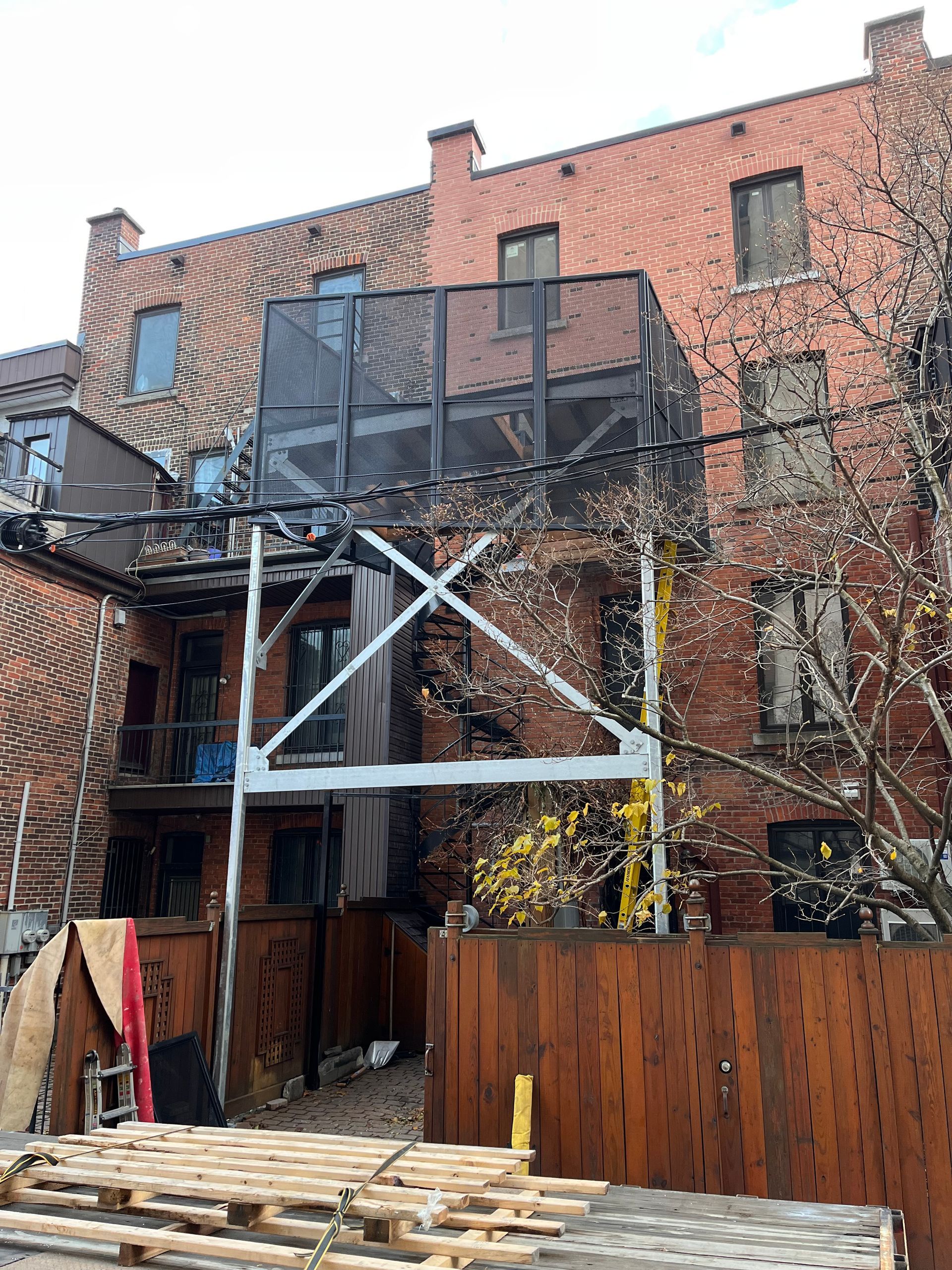 Arrière d'un bâtiment en briques avec échafaudages et plateforme clôturée. Clôture en bois et palettes au premier plan.