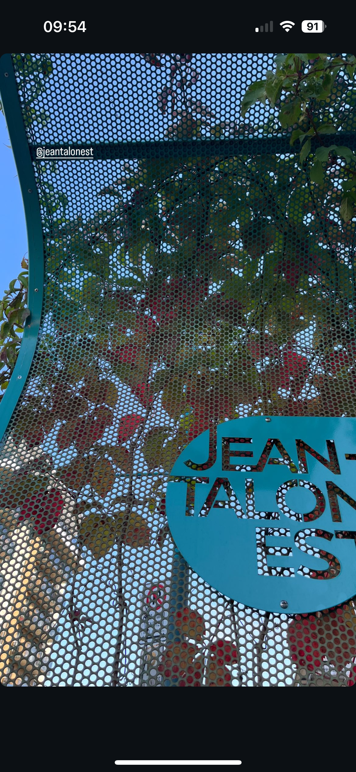Panneau indiquant le marché Jean-Talon, avec des lettres turquoise et argentées, derrière une clôture en grillage et du feuillage.