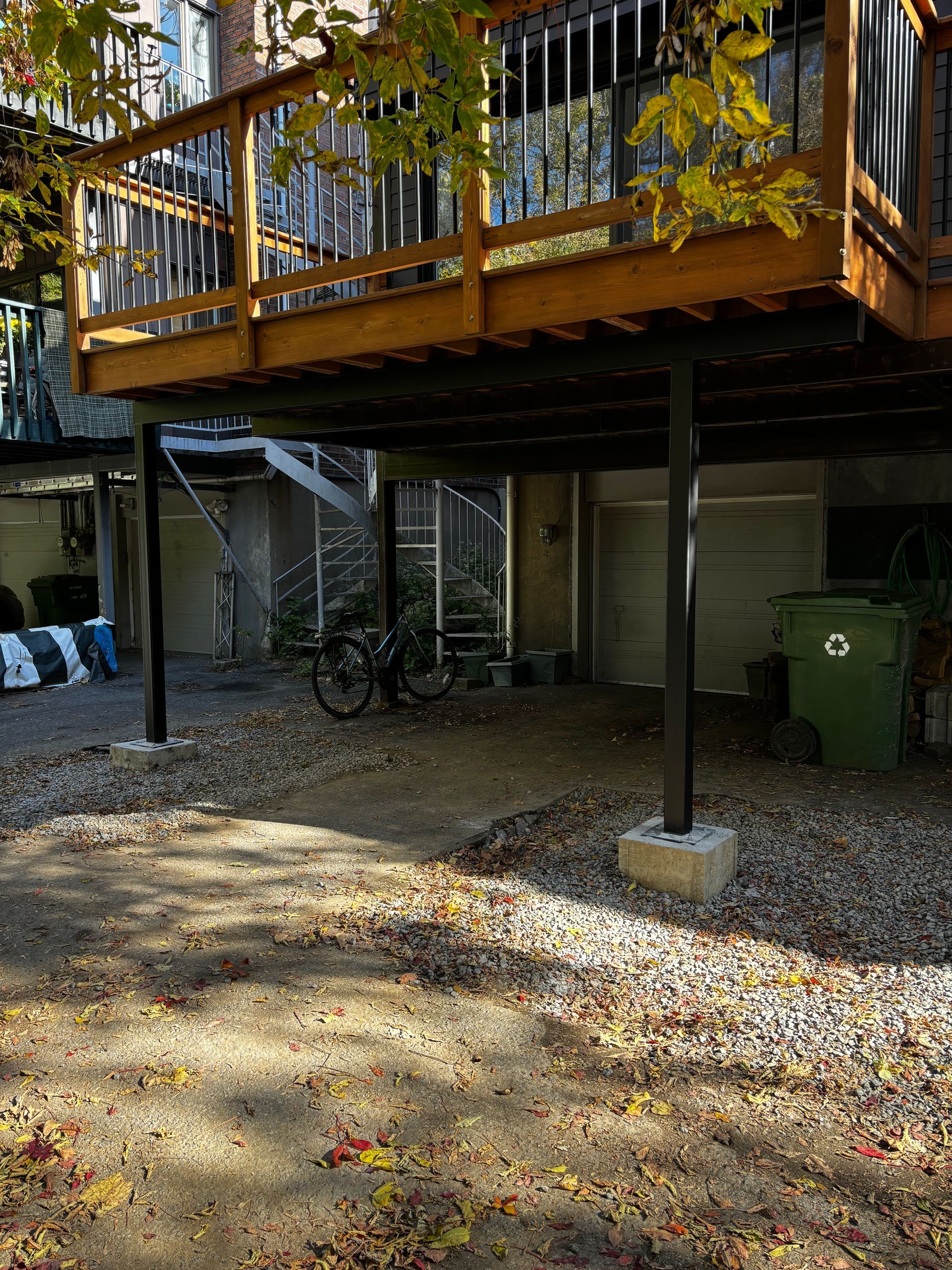 Vue d'une terrasse en bois soutenue par des poteaux noirs. En dessous, une zone gravillonnée avec un vélo, une poubelle et des portes de garage.