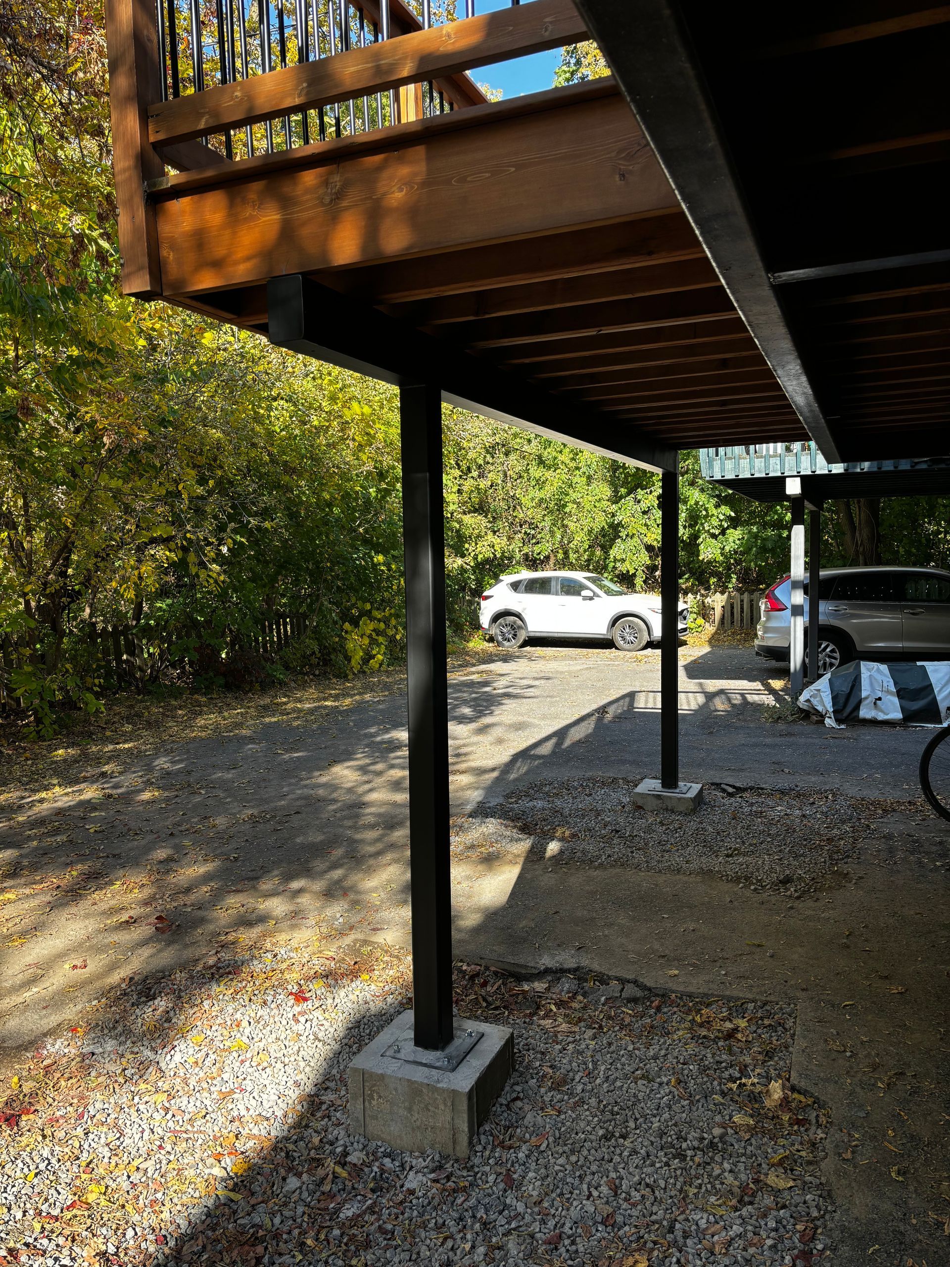 Des piliers de soutien noirs soutiennent une terrasse en bois au-dessus d'une zone de gravier où sont garées les voitures.