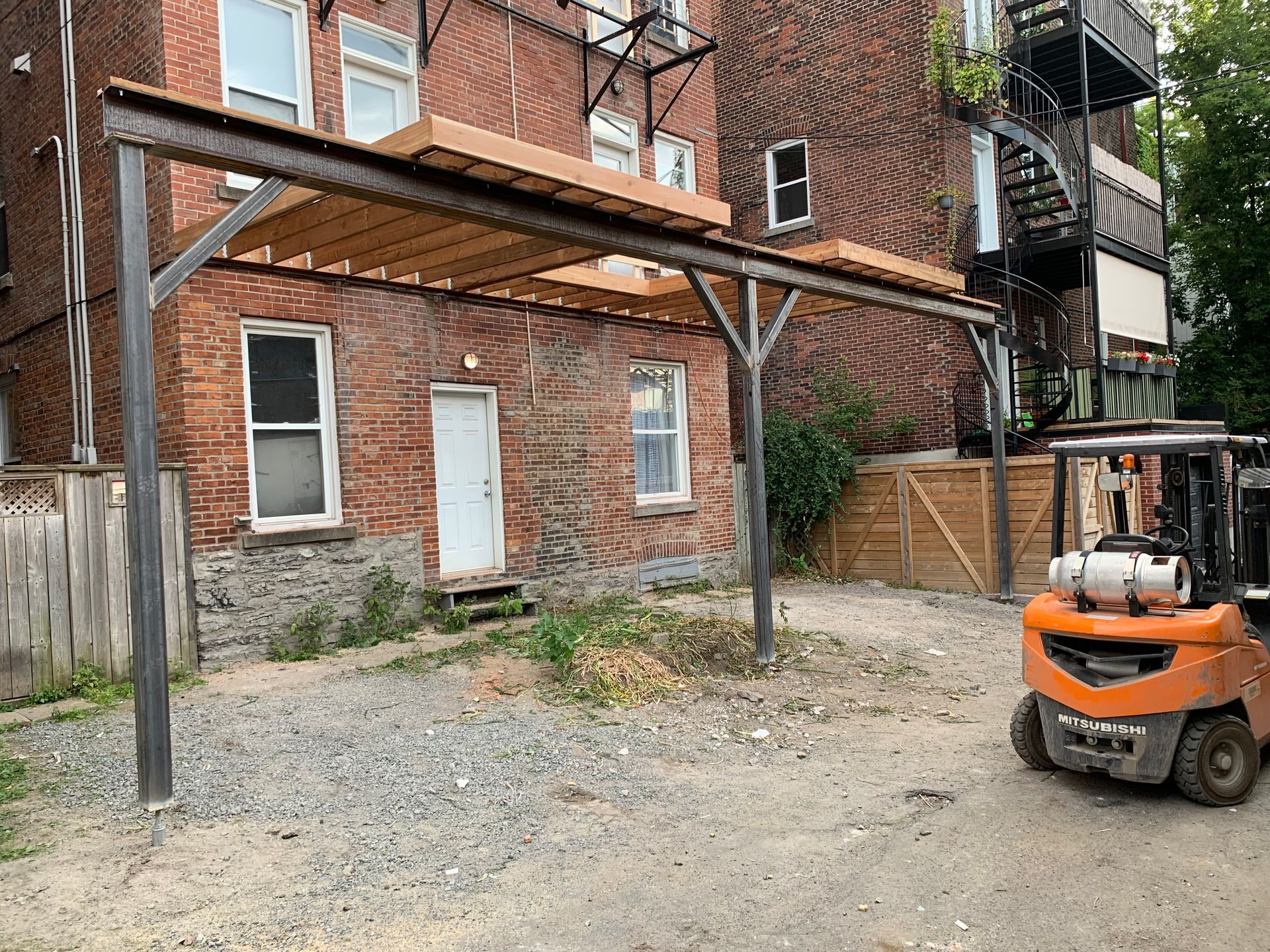 Extérieur d'un bâtiment en briques avec un auvent à structure métallique au-dessus d'une porte et de fenêtres. Un chariot élévateur est stationné sur le côté.