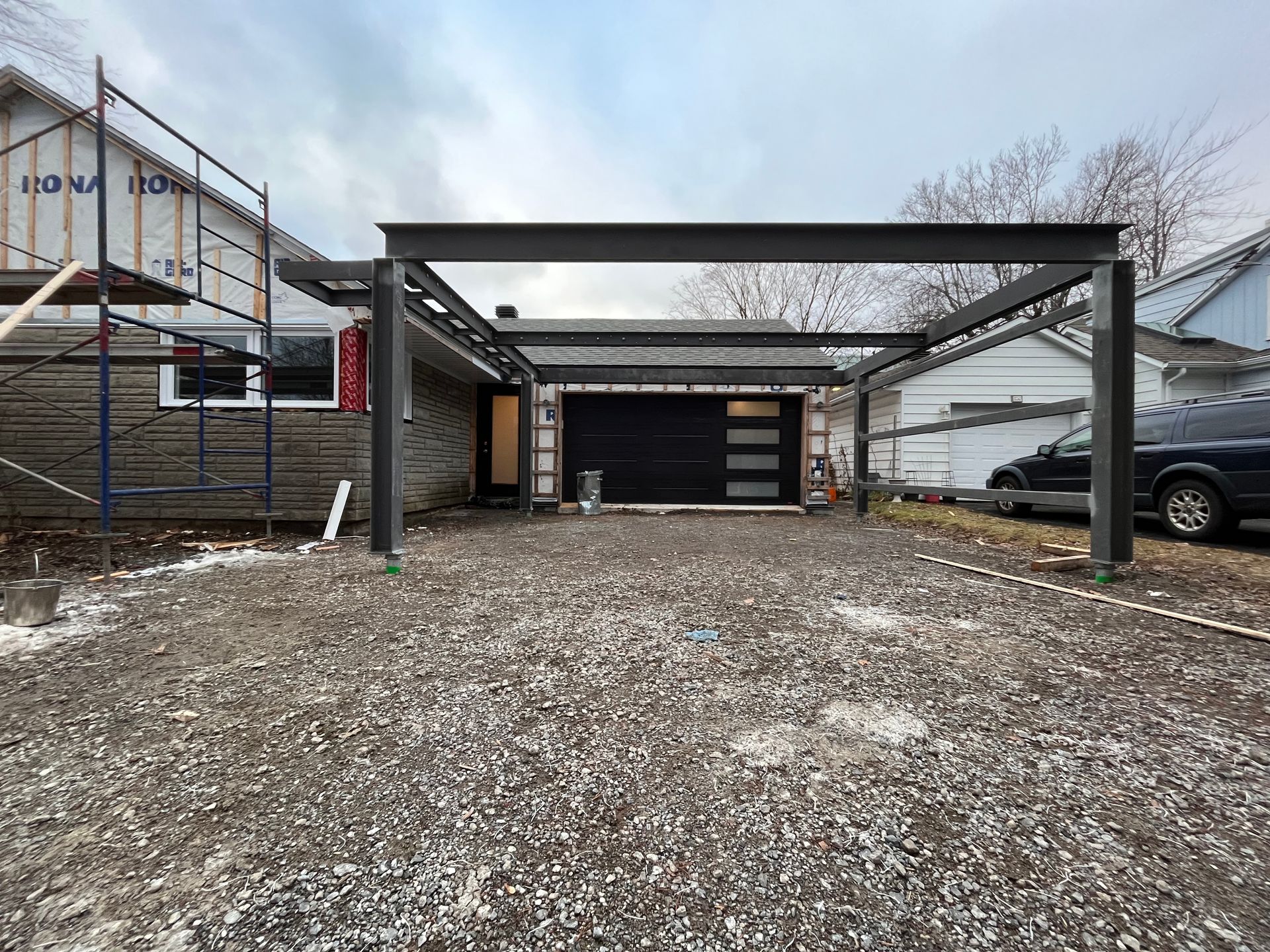 Allée avec un abri de voiture métallique au-dessus d'une porte de garage ; maison en construction à gauche, véhicules à droite.