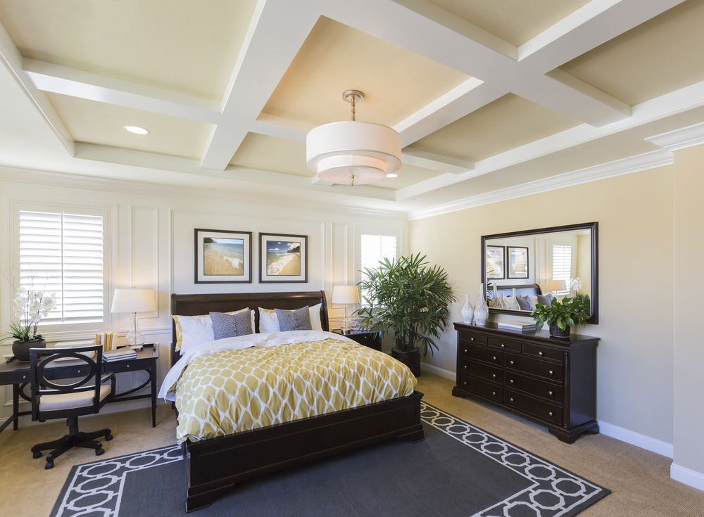 A bedroom with a dark wooden bed frame, a yellow patterned duvet, a desk with a chair, and a dresser under a mirror.