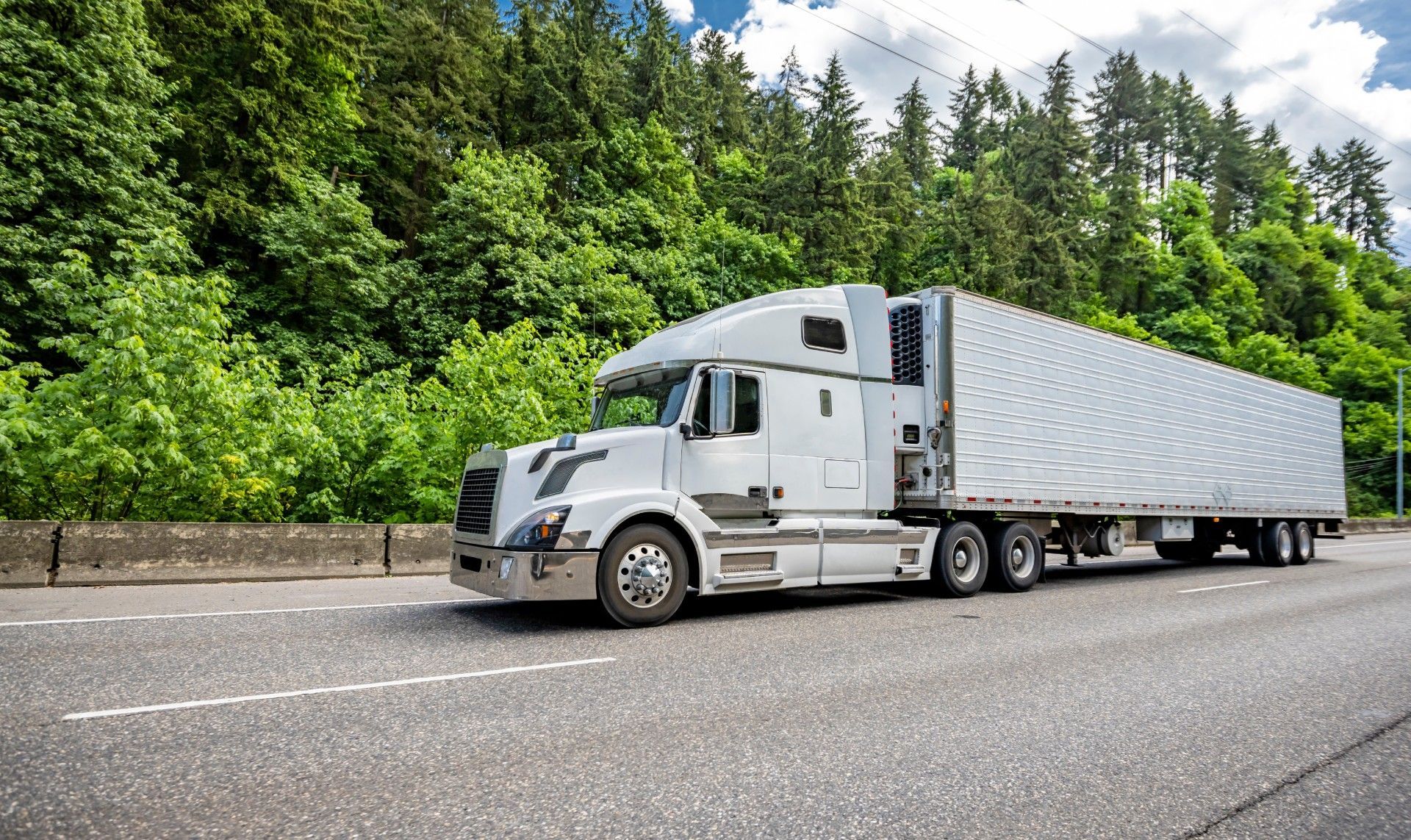 A white semi truck is driving down a highway next to a forest.