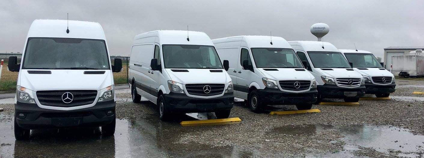 A row of white vans are parked in a parking lot on a rainy day.