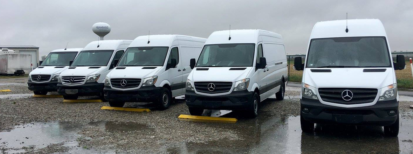 A row of white vans are parked in a parking lot on a rainy day.