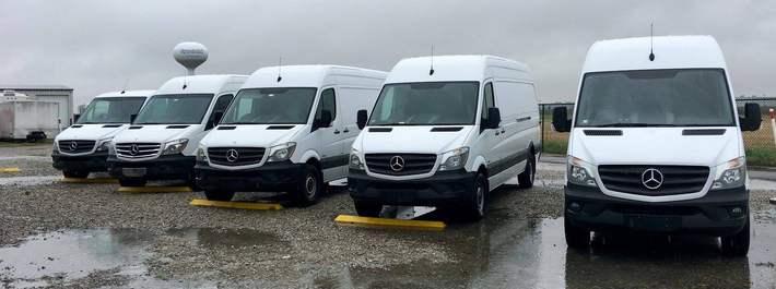 A row of white vans are parked in a parking lot on a rainy day.