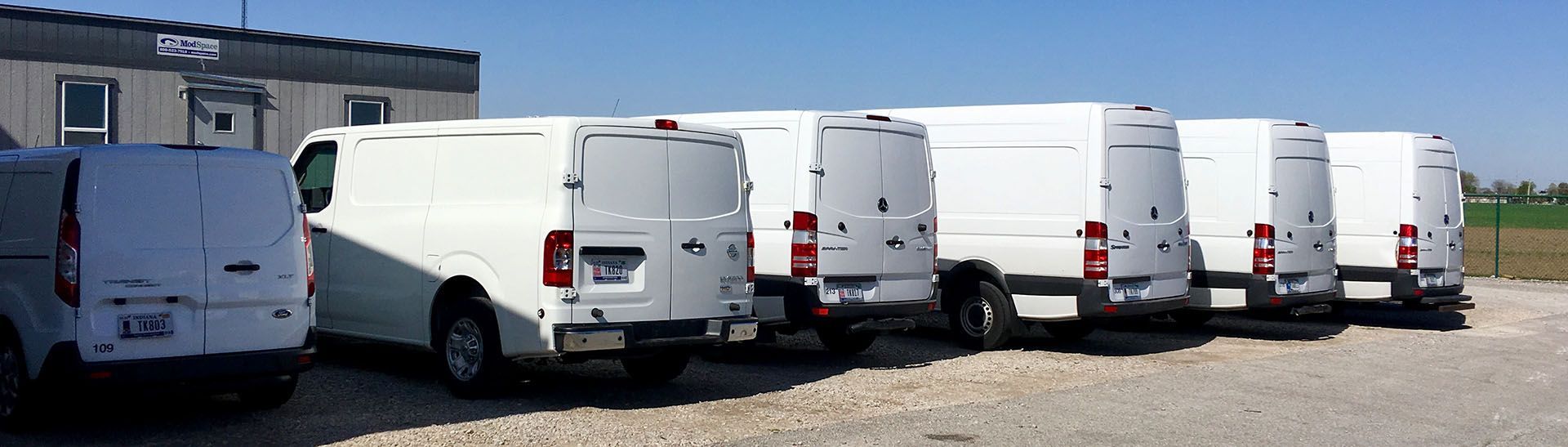 A row of white vans are parked in a gravel lot.