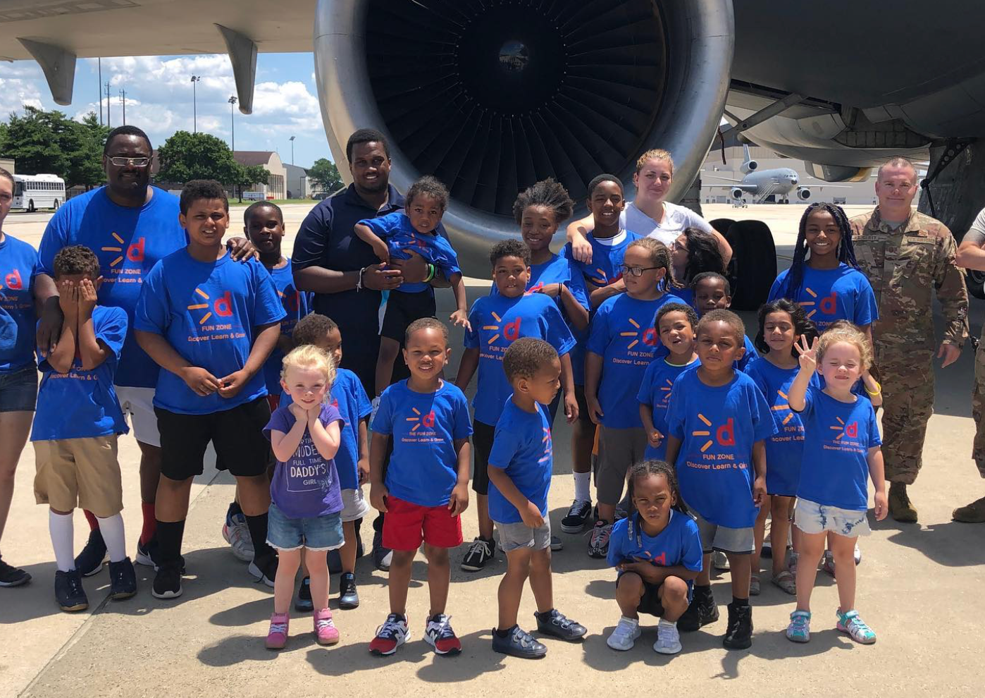 Group of children in blue shirts posing in front of a large airplane engine, with several adults.