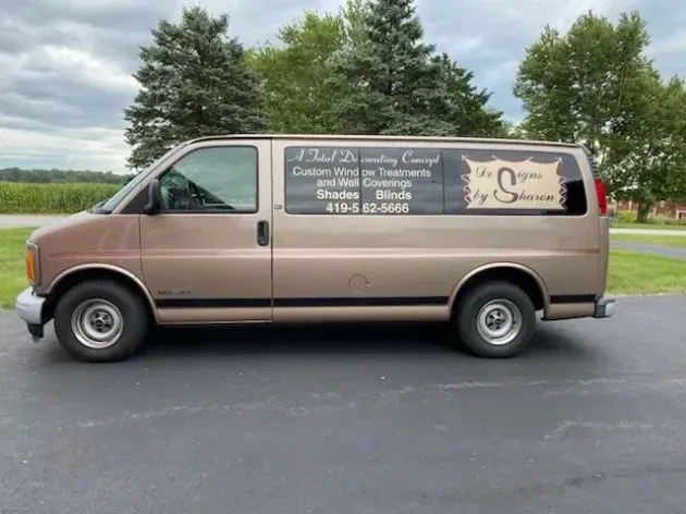 Tan cargo van with business signage parked on a road, trees in the background.