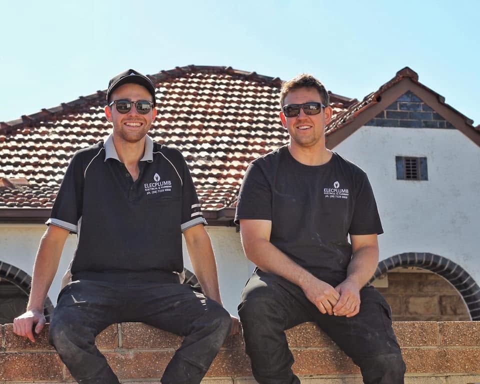 Two people in sunglasses sit on a stone wall in front of a tiled-roof building.